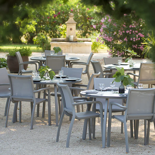 Commercial outdoor dining patio with modern Sunset sling chairs and round Sunset dining tables surrounded by lush landscaping and garden views.